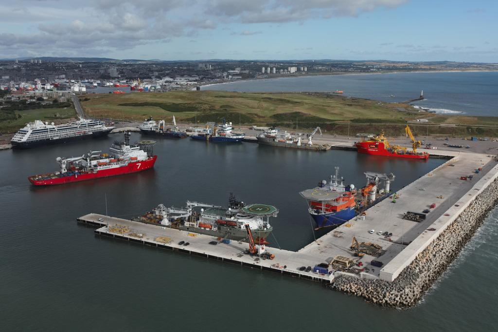 Aerial view of Port of Aberdeen's bustling South Harbour with red and blue vessels docked along its various quaysides. A large cruise ship is seen on South Harbour's Balmoral Quay, with the port's North Harbour, the Girdleness Lighthouse and Aberdeen City Centre in the distance under a partly cloudy sky.